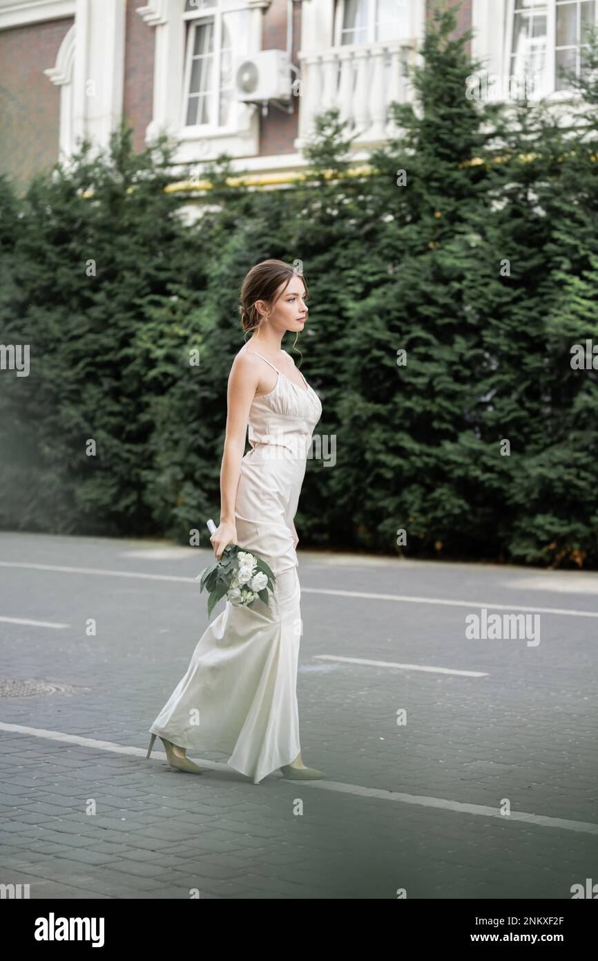 Lange junge Frau in weißem Kleid, die einen Blumenstrauß hält und draußen steht, Stockbild Stockfoto