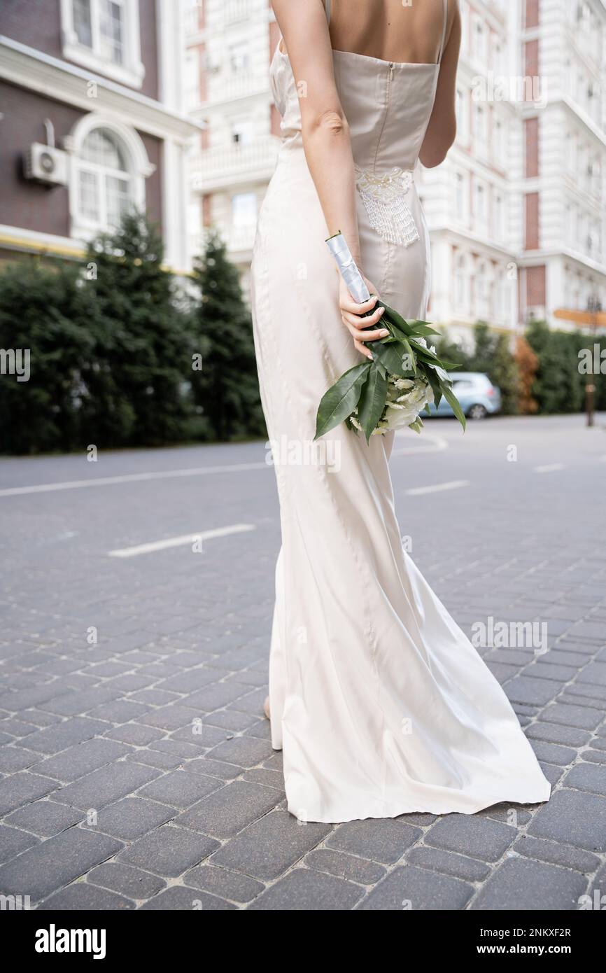 Teilansicht einer jungen Frau in weißem Kleid mit Hochzeitsstrauß hinter dem Rücken, Stockbild Stockfoto