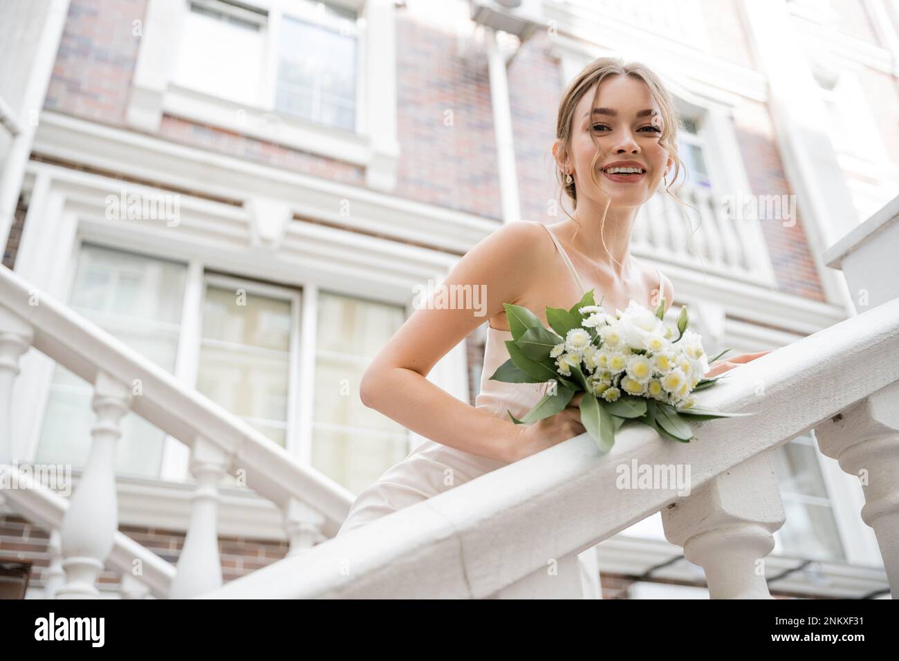 Blick auf die glückliche Braut im Hochzeitskleid mit Blumenstrauß und Blick auf die Kamera in der Nähe des Hauses, Stockbild Stockfoto