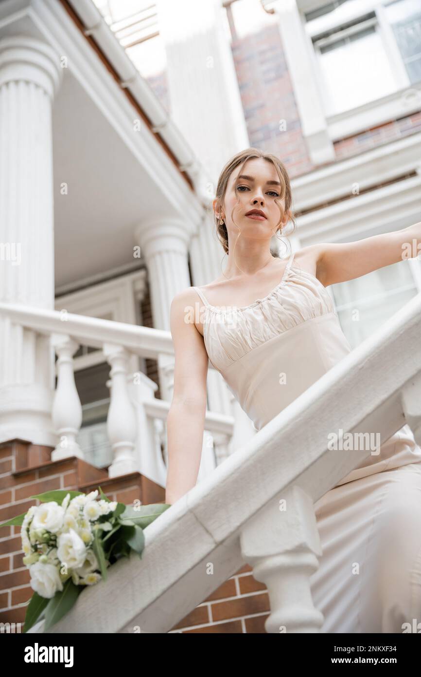 Kleiner Winkel der jungen Braut im Hochzeitskleid, die in der Nähe des Hauses auf die Kamera schaut, Stockbild Stockfoto