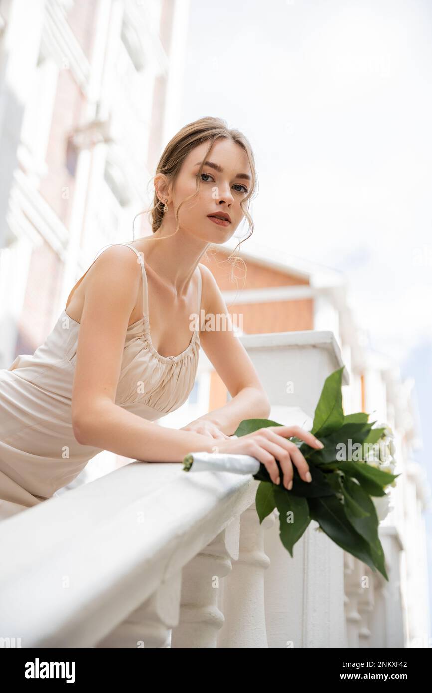 Blick aus dem niedrigen Winkel auf die wunderschöne Braut im Hochzeitskleid mit dem Blumenstrauß und Blick auf die Kamera in der Nähe des Hauses, Stockbild Stockfoto