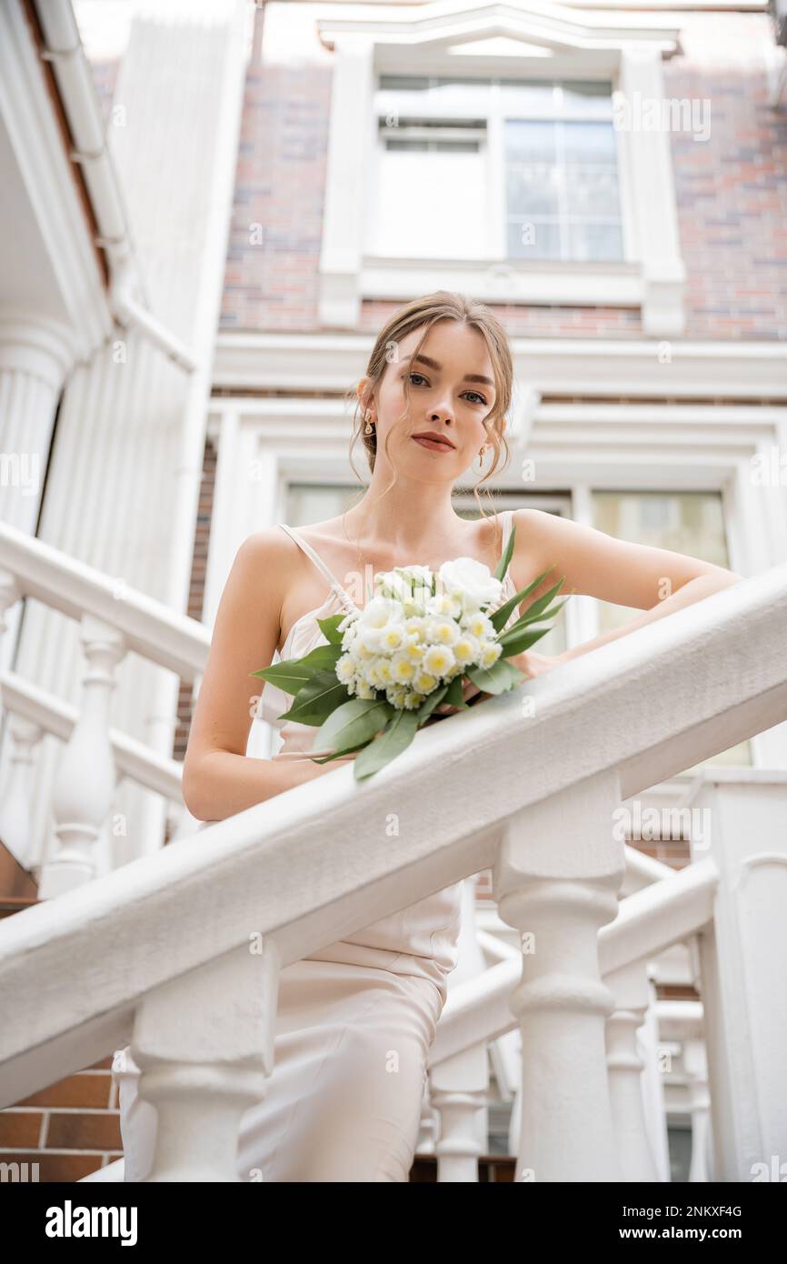 Kleiner Winkel der Braut in weißem Kleid mit Strauß und Blick auf die Kamera in der Nähe des Hauses, Stockbild Stockfoto