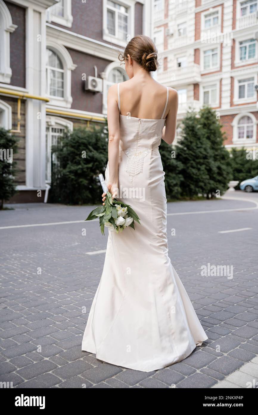 Rückansicht einer jungen Frau in weißem Kleid mit Hochzeitsstrauß hinter dem Rücken, Stockbild Stockfoto