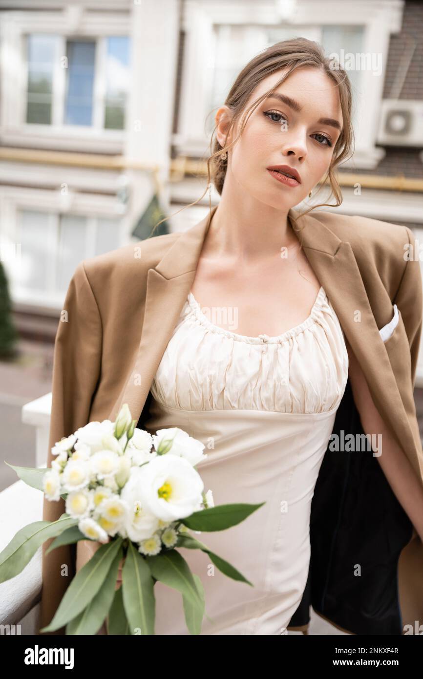 Junge Braut in Hochzeitskleid und beigefarbenem Blazer mit Blumenstrauß und Blick in die Kamera, Stockbild Stockfoto