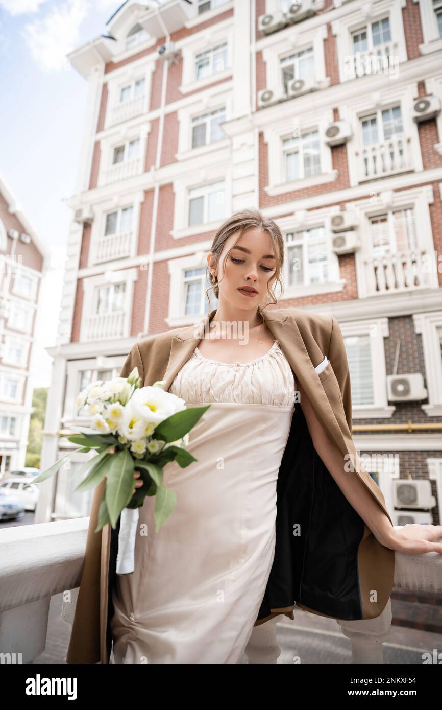 Junge Frau in Hochzeitskleid und beigefarbenem Blazer mit Blumenstrauß, Stockbild Stockfoto