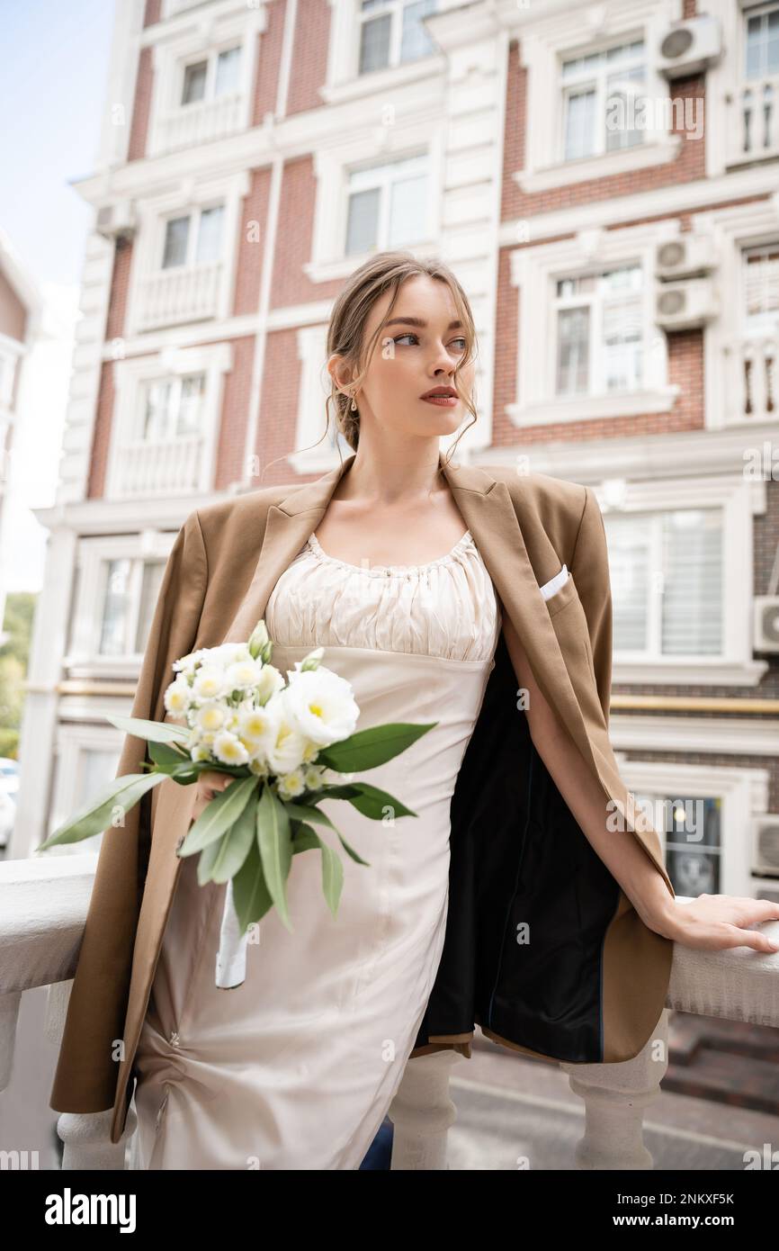 Hübsche junge Frau in Hochzeitskleid und beigefarbenem Blazer mit Blumenstrauß, Stockbild Stockfoto
