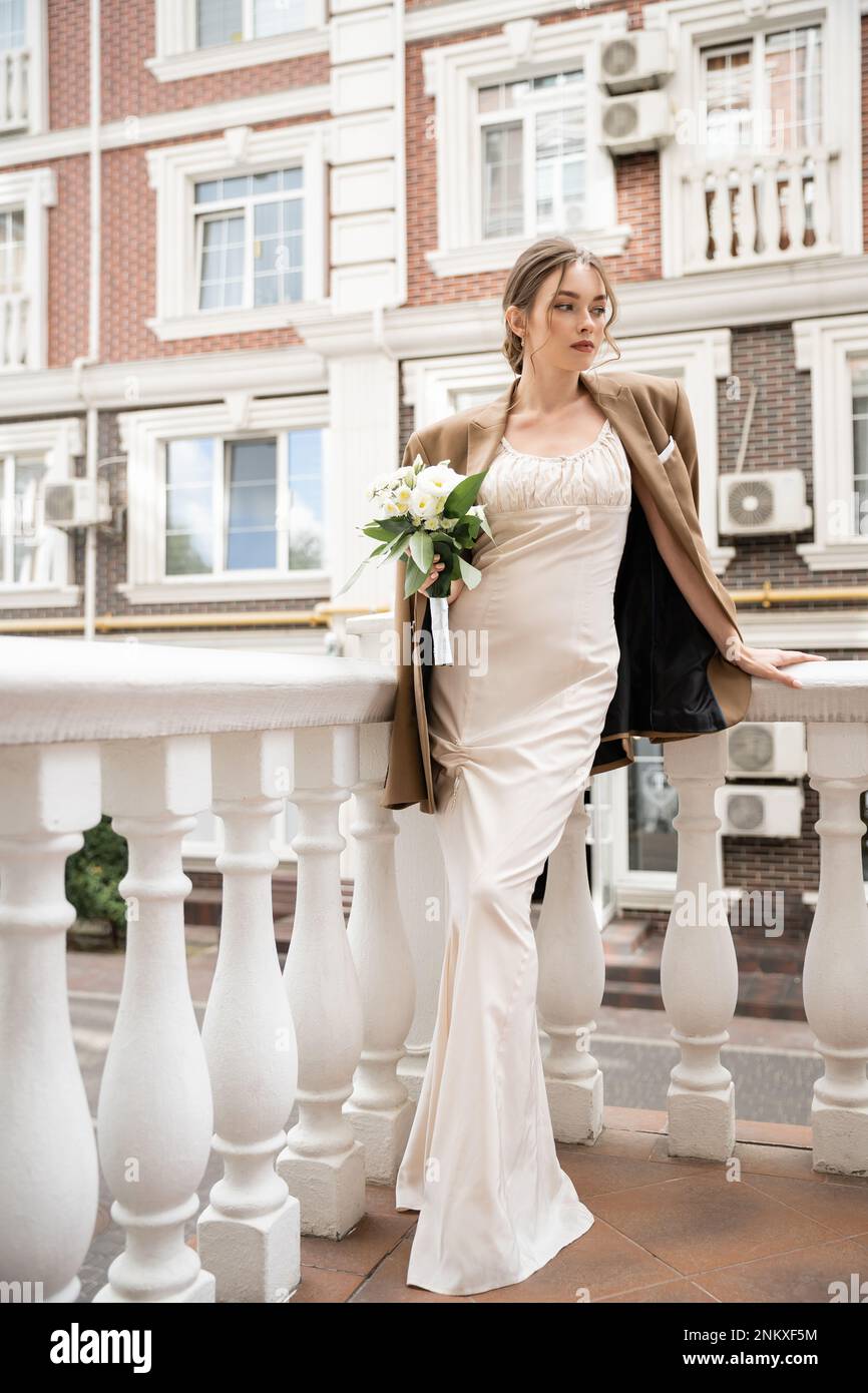 Lange Braut in Hochzeitskleid und beigefarbener Blazer mit Blumenstrauß in der Nähe des Hauses, Bild der Aktie Stockfoto