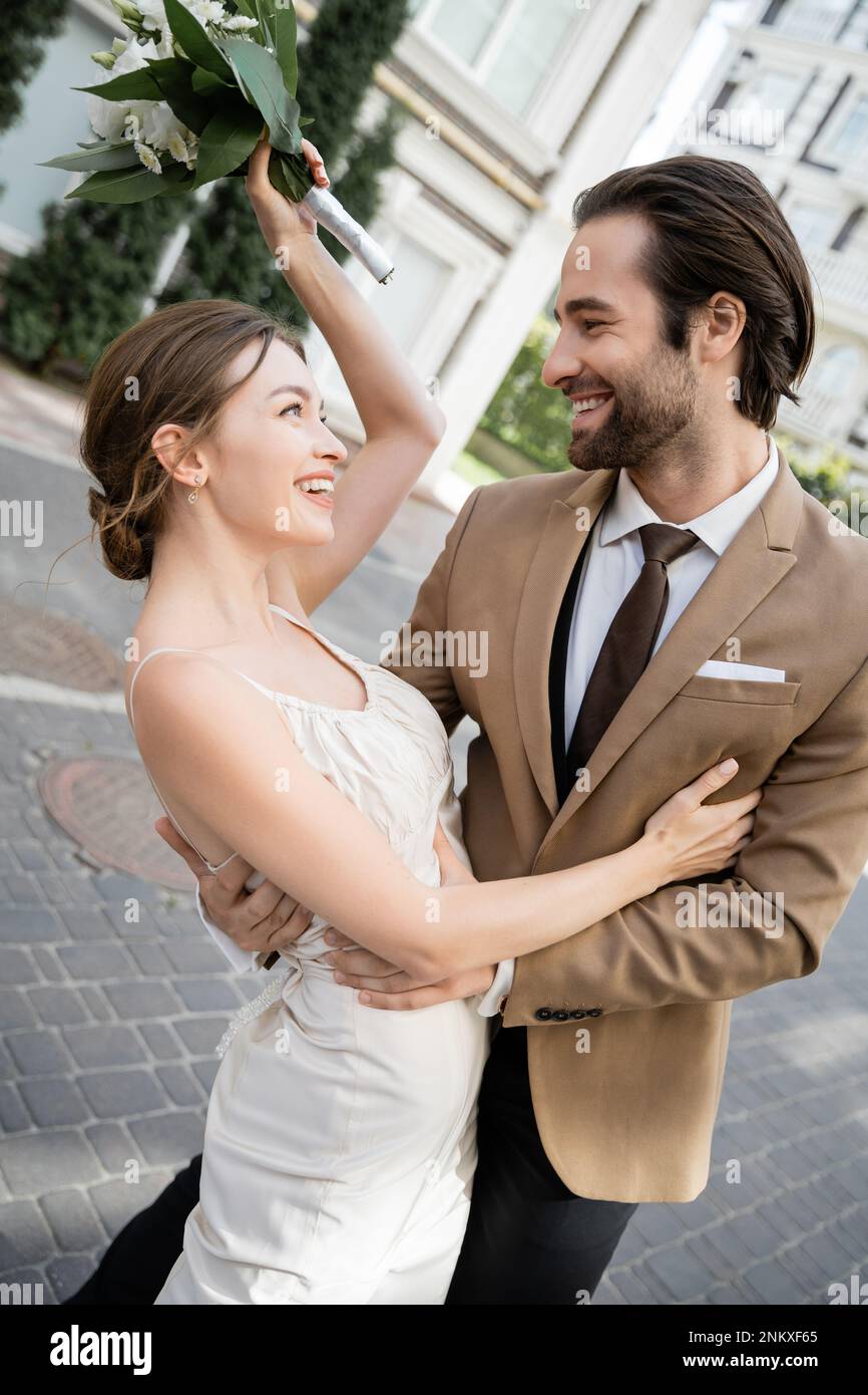Fröhliche Braut im Hochzeitskleid mit Blumenstrauß und lächelndem Bräutigam, Stockbild Stockfoto