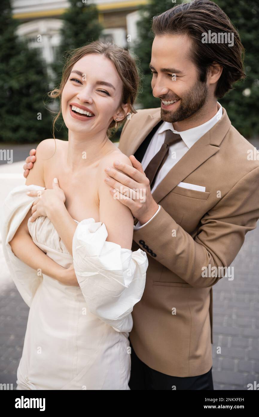 Porträt eines fröhlichen und bärtigen Mannes, der eine positive Braut in Hochzeitskleid umarmt, Stockbild Stockfoto