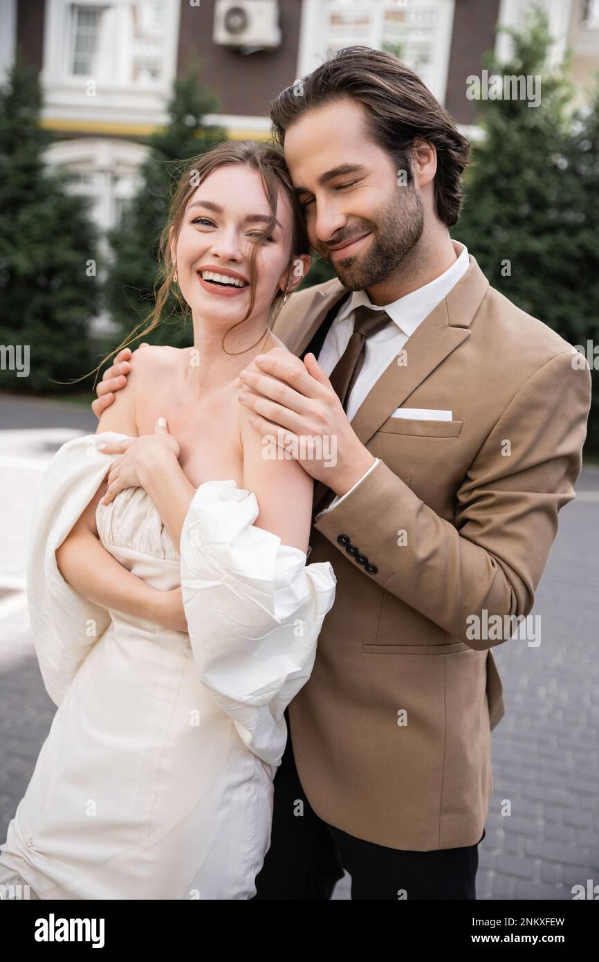 Porträt eines positiven und bärtigen Mannes, der die glückliche Braut in Hochzeitskleid umarmt, Stockbild Stockfoto