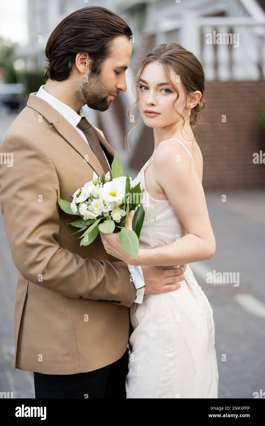 Bärtiger Bräutigam umarmte Braut in Hochzeitskleid mit blühenden Blumen, Stockbild Stockfoto