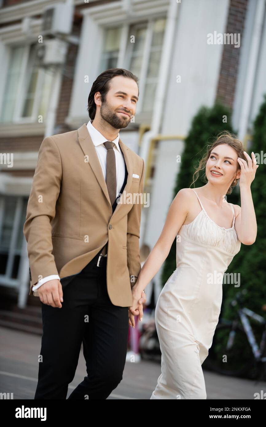 Glückliche Braut im Hochzeitskleid, die den Bräutigam ansieht, während sie Hände an der Straße hält, Stockbild Stockfoto