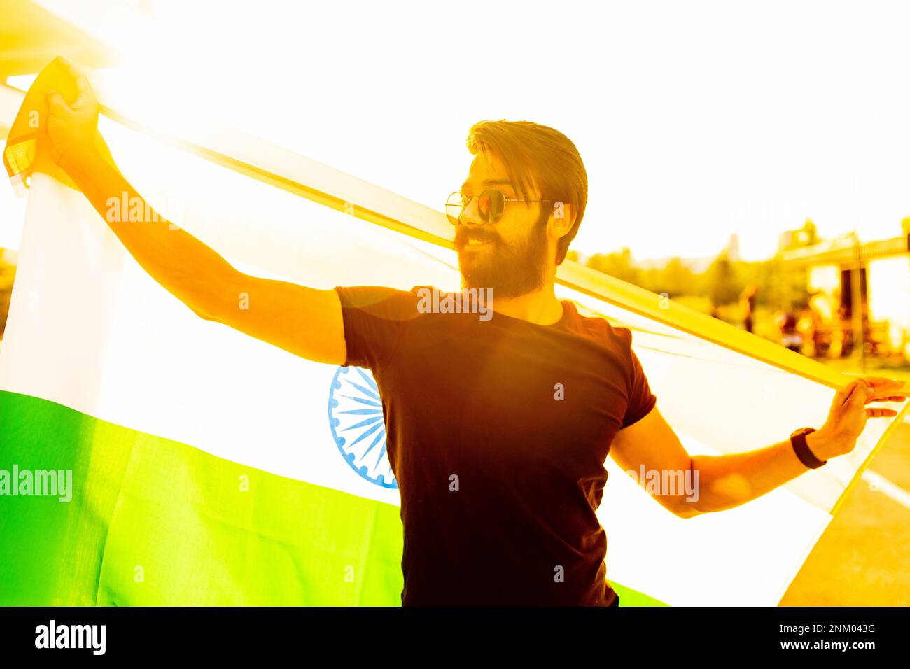 Hübscher indischer bärtiger Mann in schwarzem Baumwoll-T-Shirt, der im Sommerpark bei Sonnenuntergang bei Golglicht die indische Flagge weckte Stockfoto