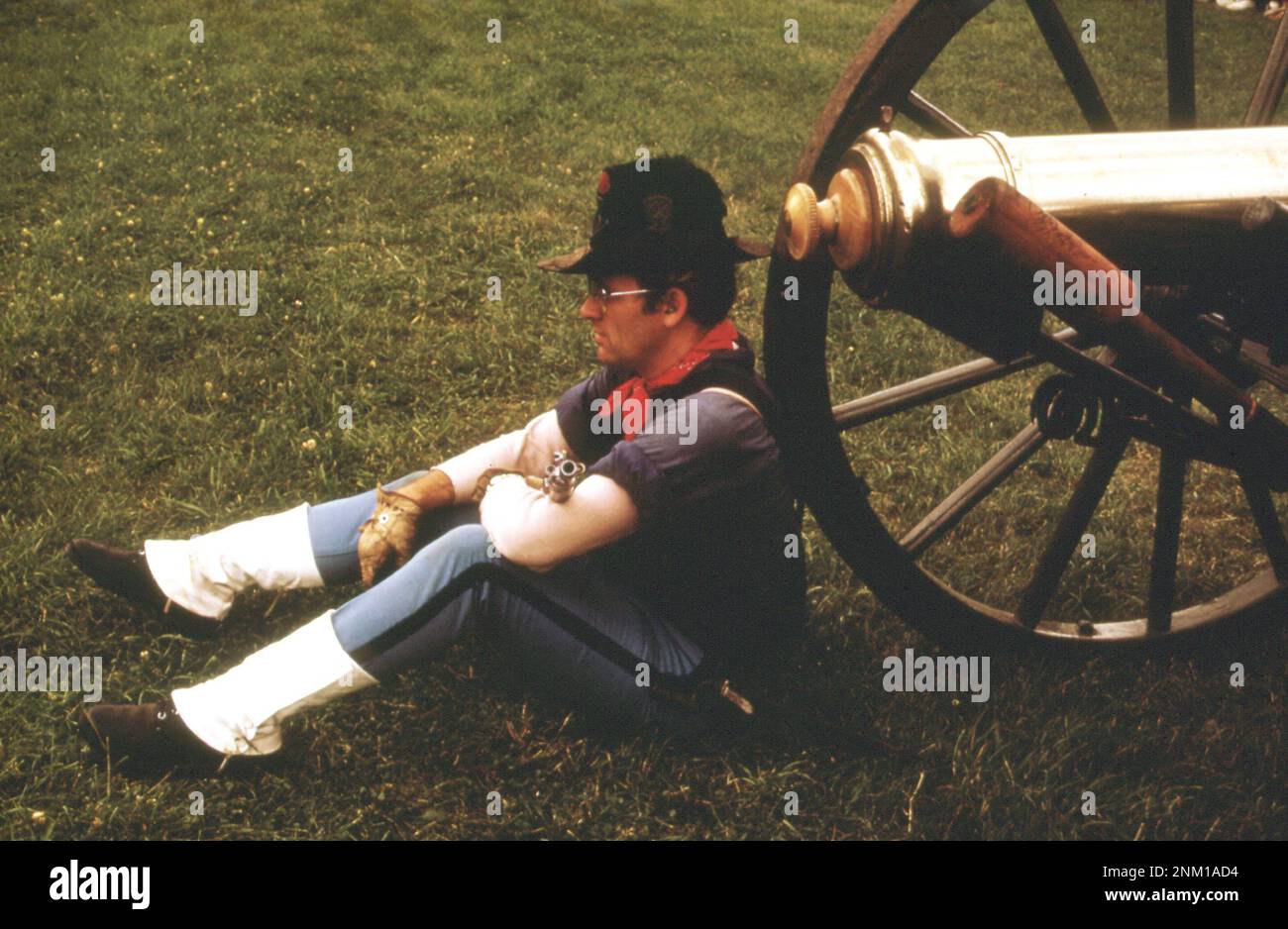 1970er America: Old Time rifleman's Meeting, 24. Juni 1973, findet auf dem Greenfield Village Green neben dem Henry Ford Museum of Dearborn statt. Ein "Soldat" ruht gegen eine alte Messingkanone Stockfoto