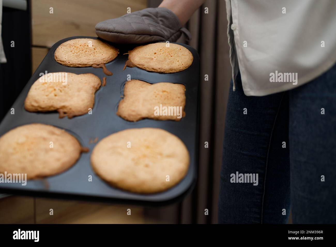 Eine Frau, die ein heißes Tablett mit gebackenen Cupcakes, Muffins und Keksen in Ofenhandschuhen hält. Person ohne Gesicht steht neben dem Ofen in der modernen Küche. Hausgemachte Küche Stockfoto