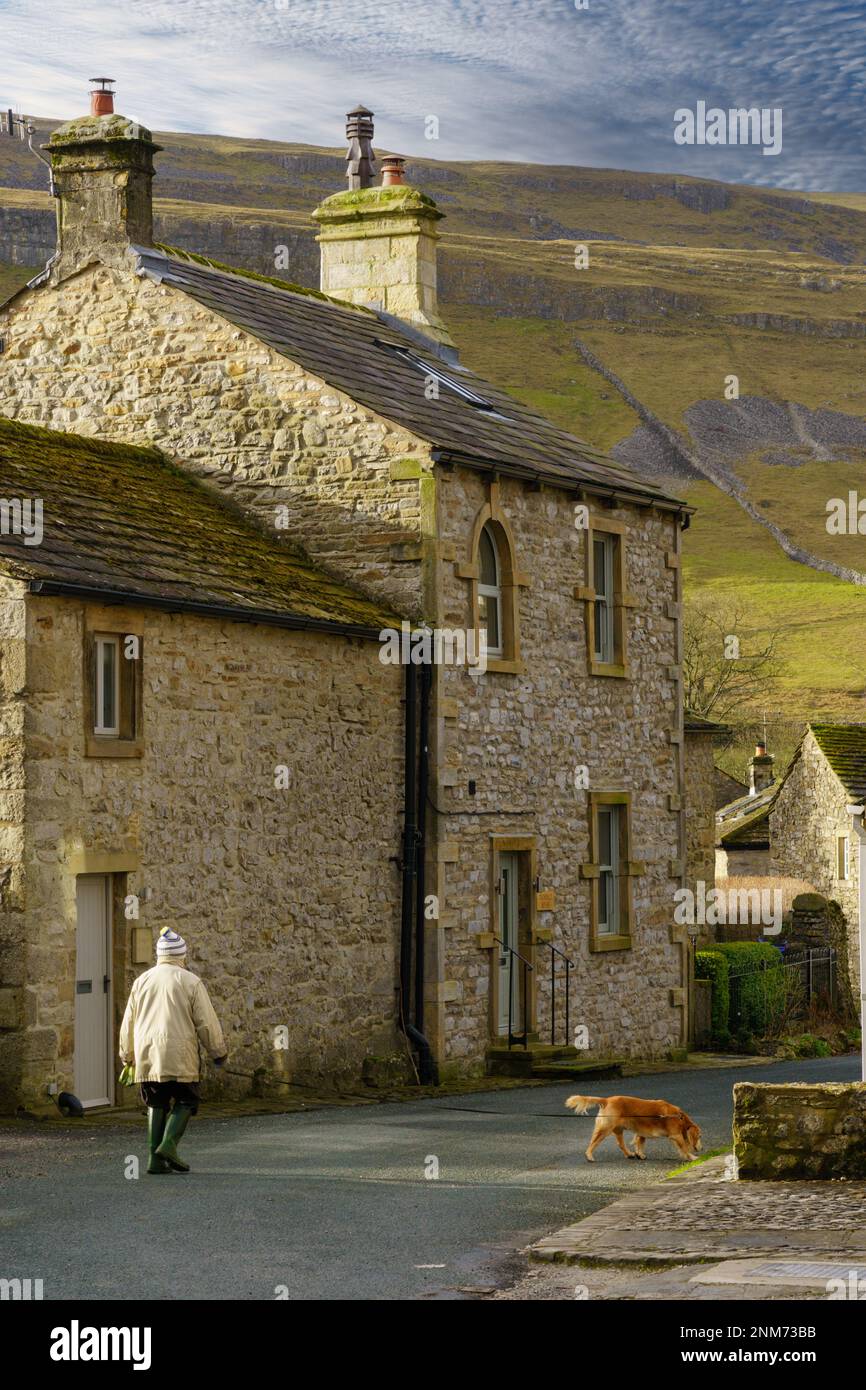An einem sonnigen Morgen führt eine Frau ihren Hund an einem rustikalen Steinhaus mit sanften Hügeln in der Ferne vorbei, Upper Wharfedale, North Yorkshire, Großbritannien. Stockfoto