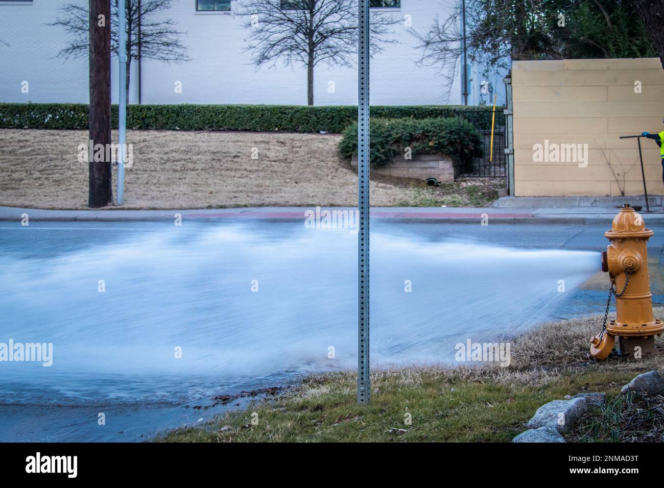 Gelber Hydrant an der Ecke der Kreuzung, der Wasser über die Straße sprudelt, und Mann auf der anderen Seite mit Werkzeug, das sich im Schachtloch auf dem Bürgersteig dreht. Stockfoto