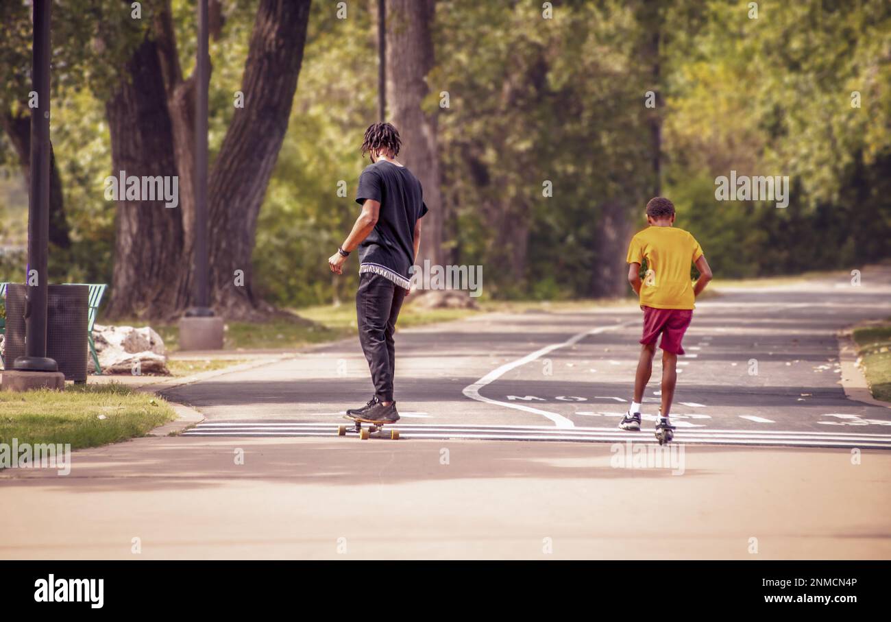 Zwei schwarze Jungs, einer auf dem Roller und einer auf dem Skateboard auf dem Parkweg, mit hohen Bäumen auf einer Seite - Bewegungsunschärfe und von der Kamera weggerichtet Stockfoto