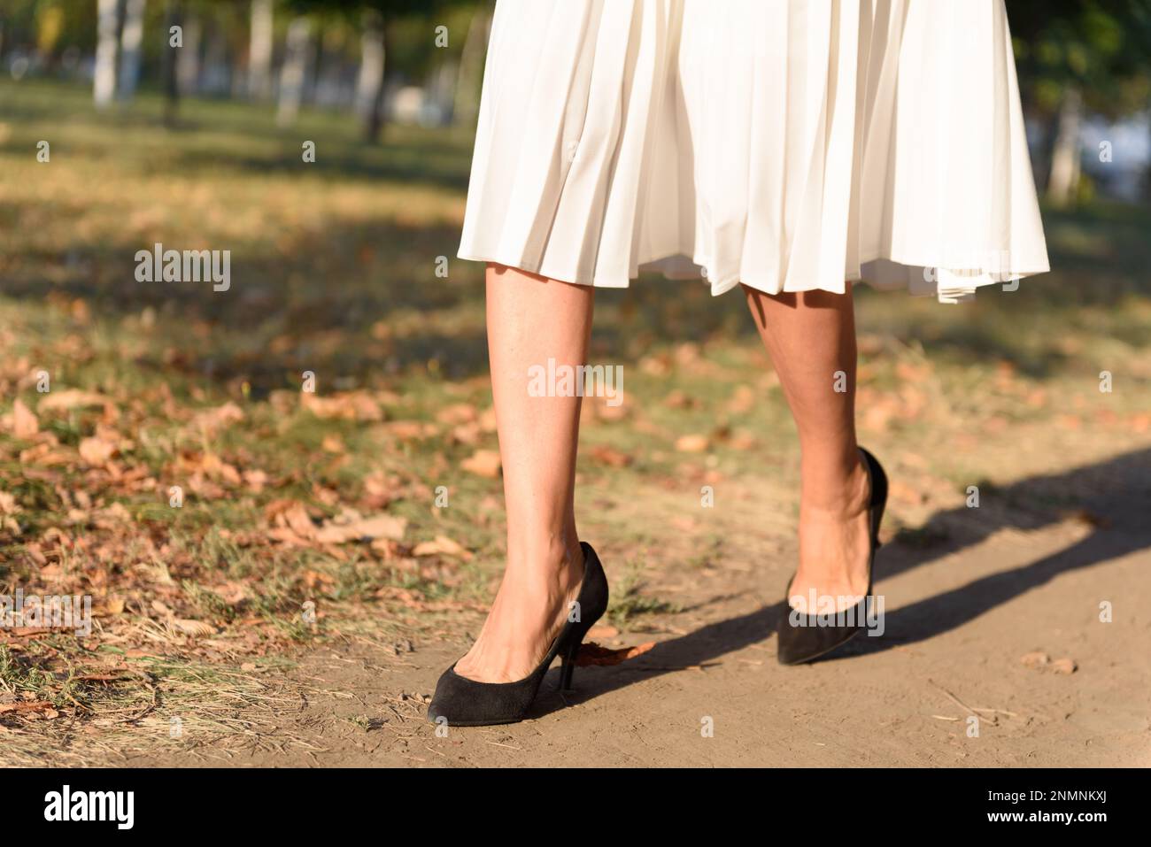 Eine Frau mit weißem Faltenrock und schwarzen Wildlederstilettos, die nach der Arbeit im Herbstpark spaziert. Stockfoto