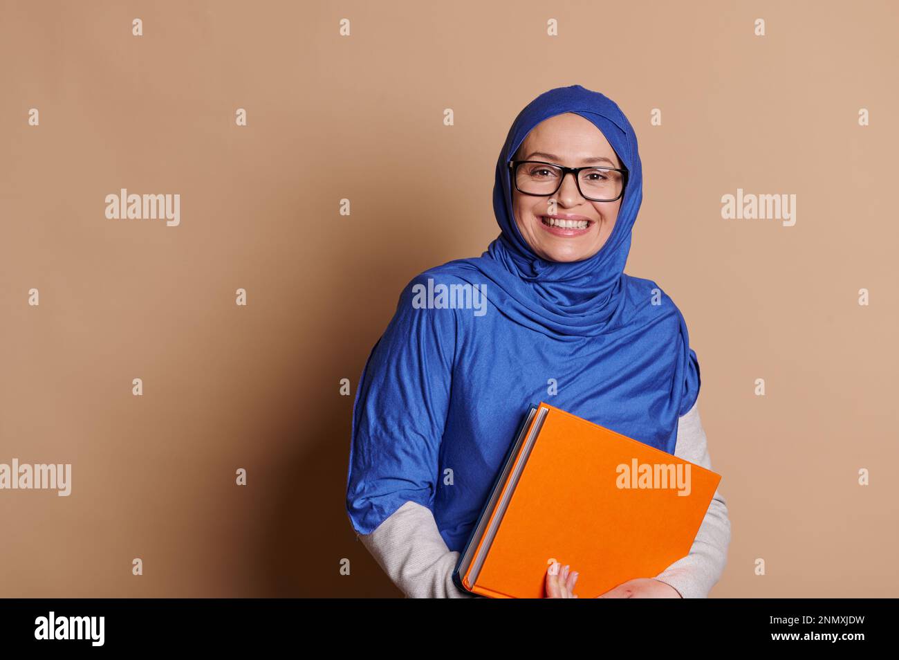 Positive, reife Lehrerin in blauem Hijab, lächelt ein zahniges Lächeln, schaut in die Kamera, hält Bücher, beiger Hintergrund Stockfoto