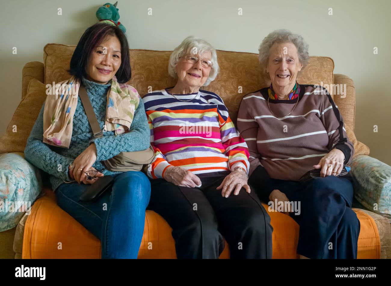 Zwei sehr ältere englische Frauen im Alter von 90s Jahren sitzen auf einem Sofa mit einer vietnamesischen Frau mittleren Alters. Stockfoto