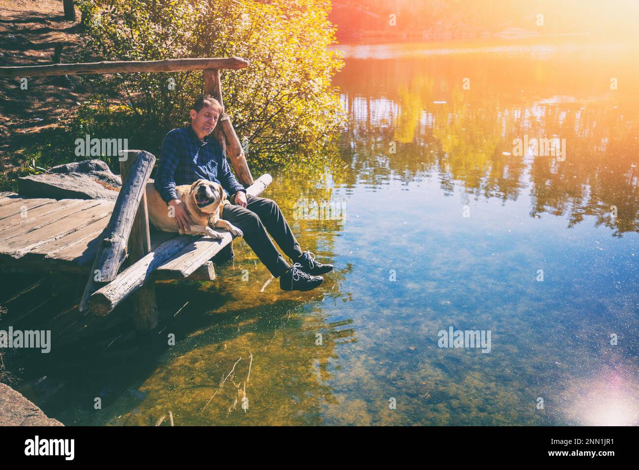 Mann mit Labrador Retriever Hund, der auf einer hölzernen Terrasse am Seeufer sitzt und Wasser sieht Stockfoto