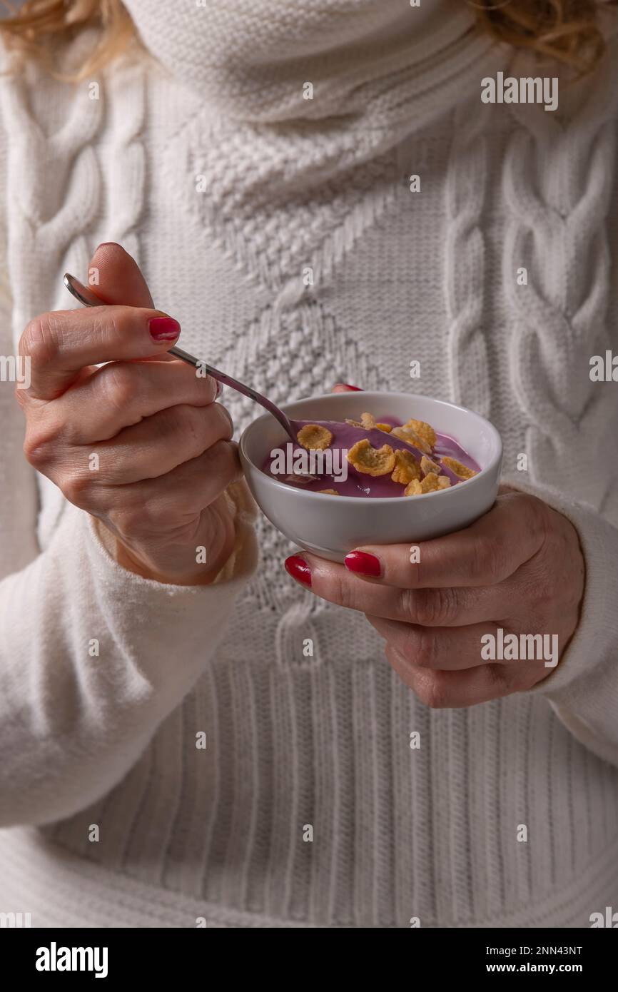 Eine Frau in einem Pullover aus weißer Wolle hält einen lila Joghurt mit Müsli drauf, den sie gleich essen wird Stockfoto