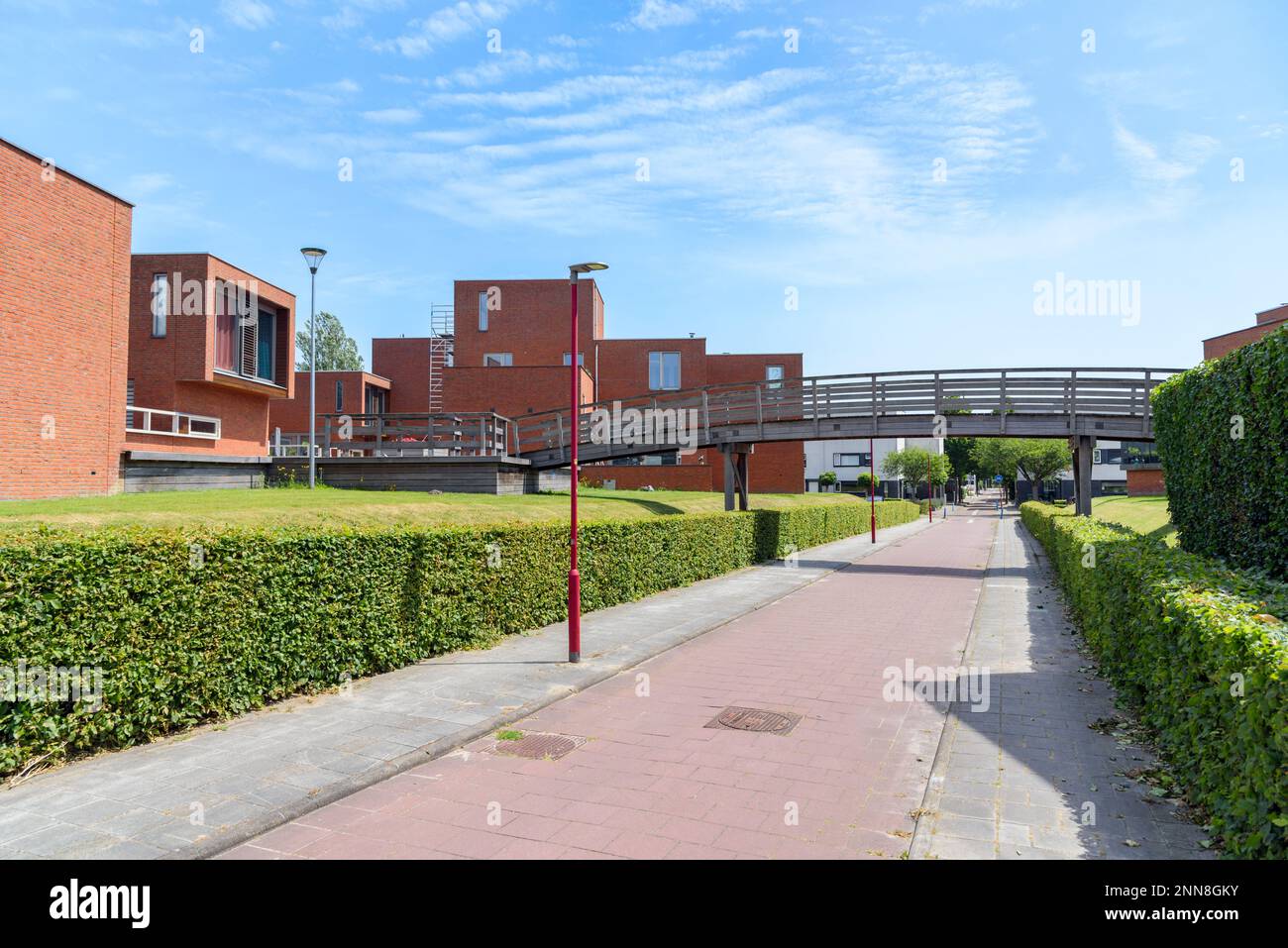 Neue Backsteinhäuser entlang eines verlassenen Fahrradwegs in einer Wohnungsbausiedlung. Eine bewaldete Fußgängerbrücke befindet sich im Vordergrund. Stockfoto