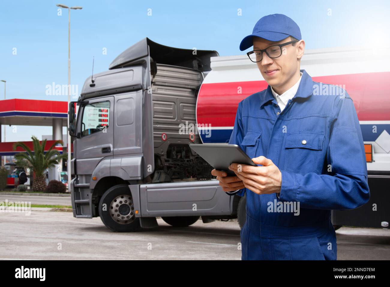 Mann mit einem digitalen Tablet auf einem Hintergrund eines Benzinlaster mit Tankwagen und einer Tankstelle Stockfoto