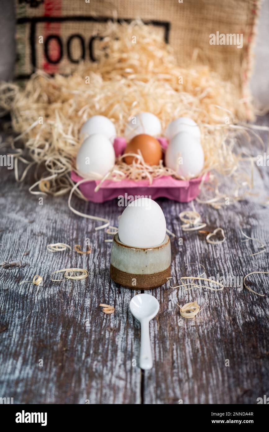 Ei in einem Eierbecher mit einem kleinen weißen Löffel, mit 6 Eiern in einer rosafarbenen Eierschachtel dahinter auf einem mit Stroh überzogenen Holzbrett Stockfoto