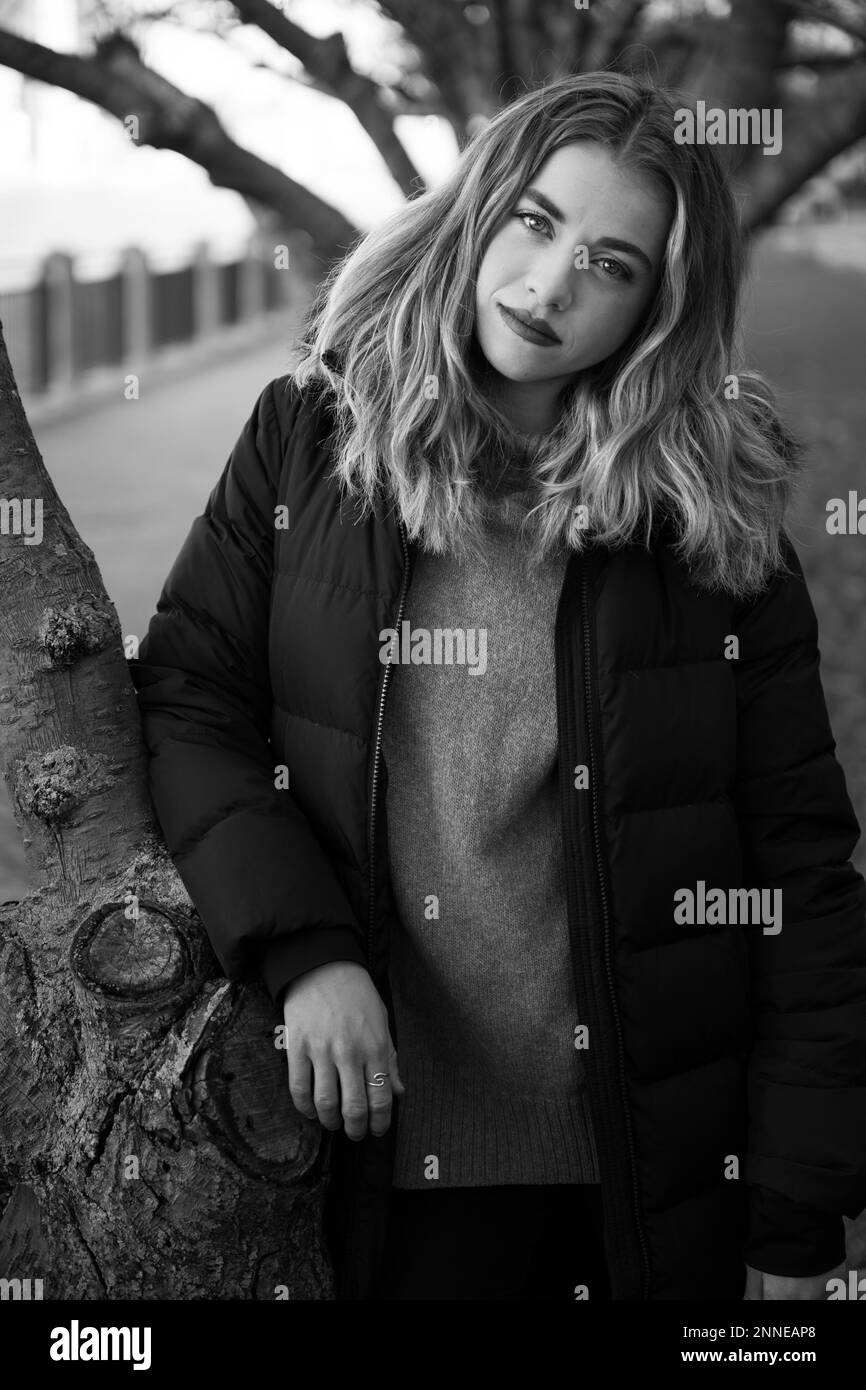 Eine graue Aufnahme einer jungen Frau mit einem ernsten Gesicht, die vor einem Baum steht und eine stilvolle Jacke trägt Stockfoto