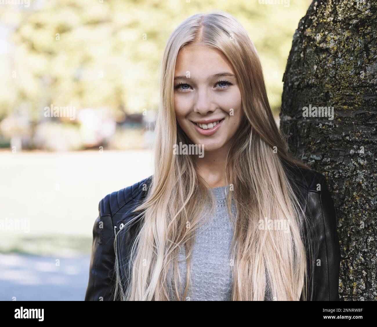 Junge Frau mit langen blonden Haaren und Lederjacke lehnte sich gegen den Baum an einem sonnigen Tag im Frühjahr Stockfoto