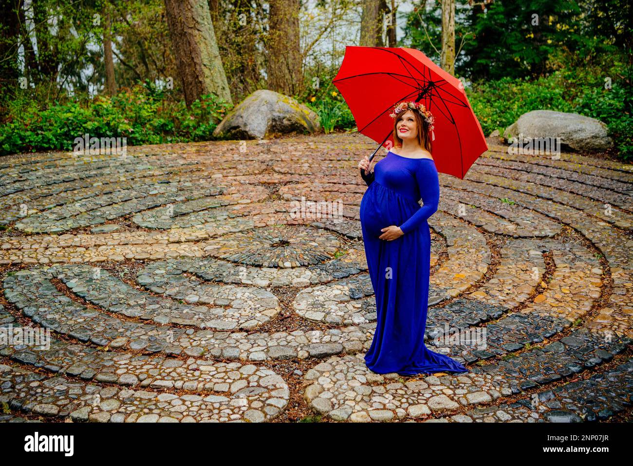 Schwangere in blauem Kleid vor einem Steinlabyrinth mit rotem Schirm Stockfoto