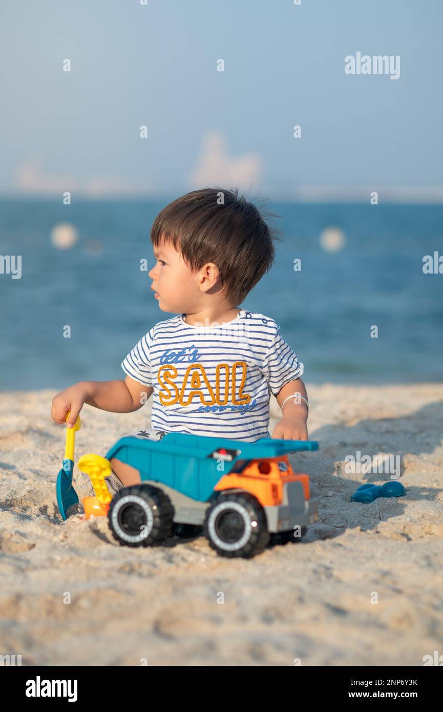 Ein Kleinkind spielt fröhlich mit einem Spielzeugfahrzeug am Strand und genießt die Wunder der Kindheit am Meer. Ein kleiner Junge, der mit einem Spielzeugtruk am Strand spielt Stockfoto