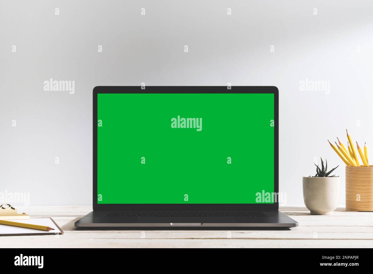 Chroma Key Laptop mit grünem Bildschirm auf dem Tisch vor dem weißen Hintergrund. Tischaufnahme des Innenraums mit Fensterlichteffekt. Notizblock, Bleistifte und Stockfoto