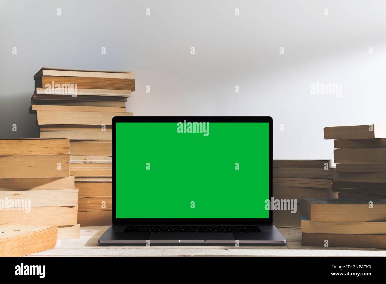 Chroma Key Laptop mit grünem Bildschirm und ein Stapel Bücher auf dem Tisch vor dem weißen Hintergrund. Tischaufnahme des Innenraums mit Fensterlichteffekt. Stockfoto