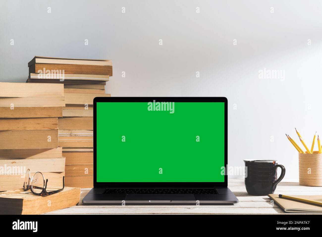 Chroma Key Laptop mit grünem Bildschirm und ein Stapel Bücher auf dem Tisch vor dem weißen Hintergrund. Tischaufnahme des Innenraums mit Fensterlichteffekt. Stockfoto