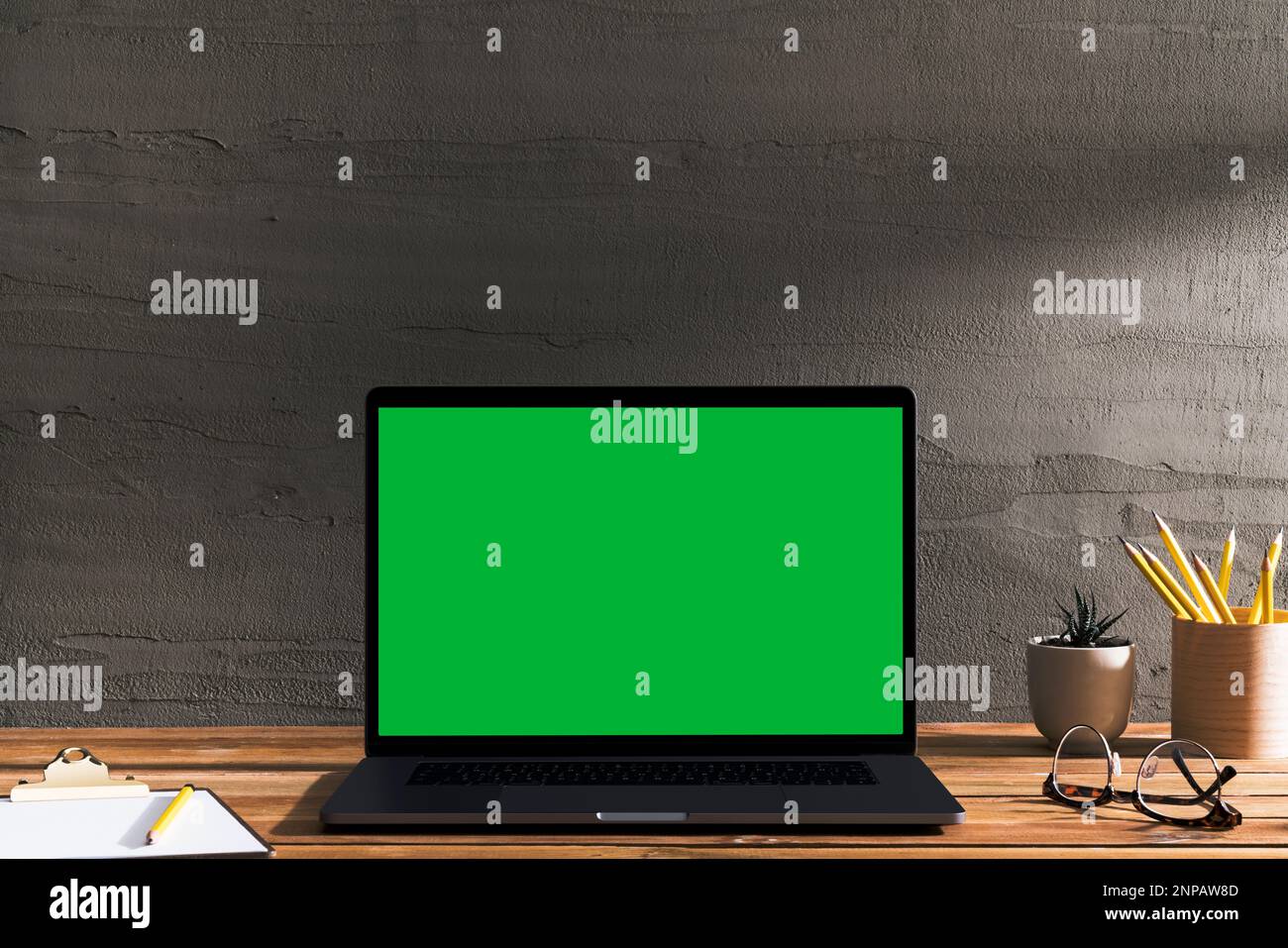 Chroma Key Laptop mit grünem Bildschirm auf Holztisch vor Betonwand. Tischaufnahme des Innenraums mit Fensterlichteffekt. Stockfoto