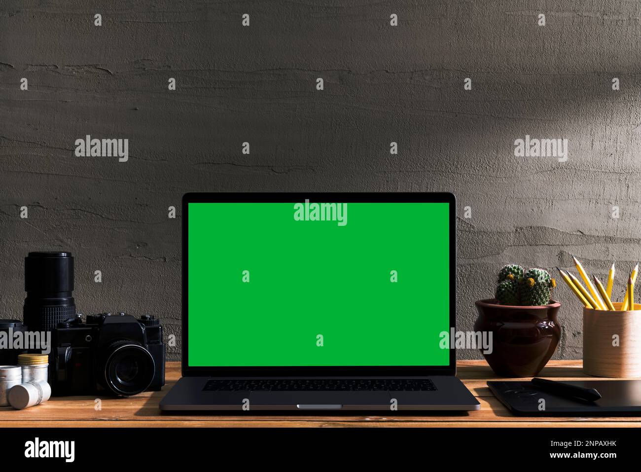 Chroma Key Laptop mit grünem Bildschirm und Fotoausrüstung auf Holztisch vor Betonwand. Tischaufnahme des Innenraums mit Fensterbeleuchtung Stockfoto