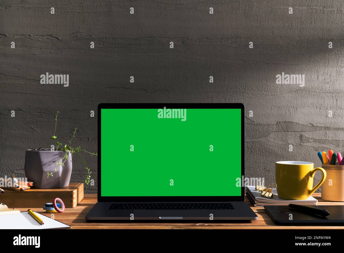 Chroma Key Laptop mit grünem Bildschirm auf Holztisch des Designers vor Betonwand. Tischaufnahme des Innenraums mit Fensterlichteffekt. Stockfoto