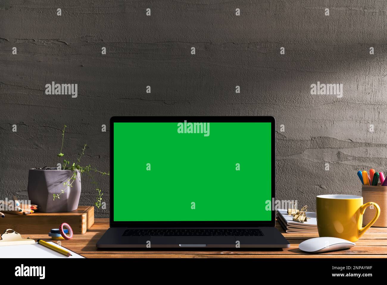 Chroma Key Laptop mit grünem Bildschirm auf Holztisch des Designers vor Betonwand. Tischaufnahme des Innenraums mit Fensterlichteffekt. Stockfoto