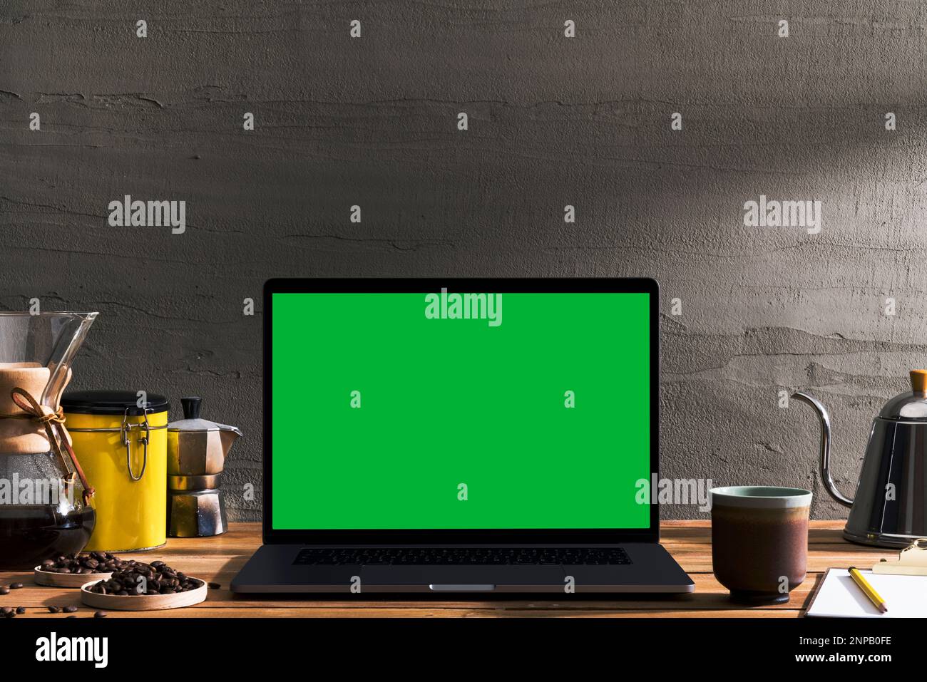 Chroma Key Laptop mit grünem Bildschirm und Werkzeuge zum Brühen von Kaffee auf einem Holztisch vor einer Betonwand. Tischaufnahme des Innenraums mit Fensterbeleuchtung Stockfoto