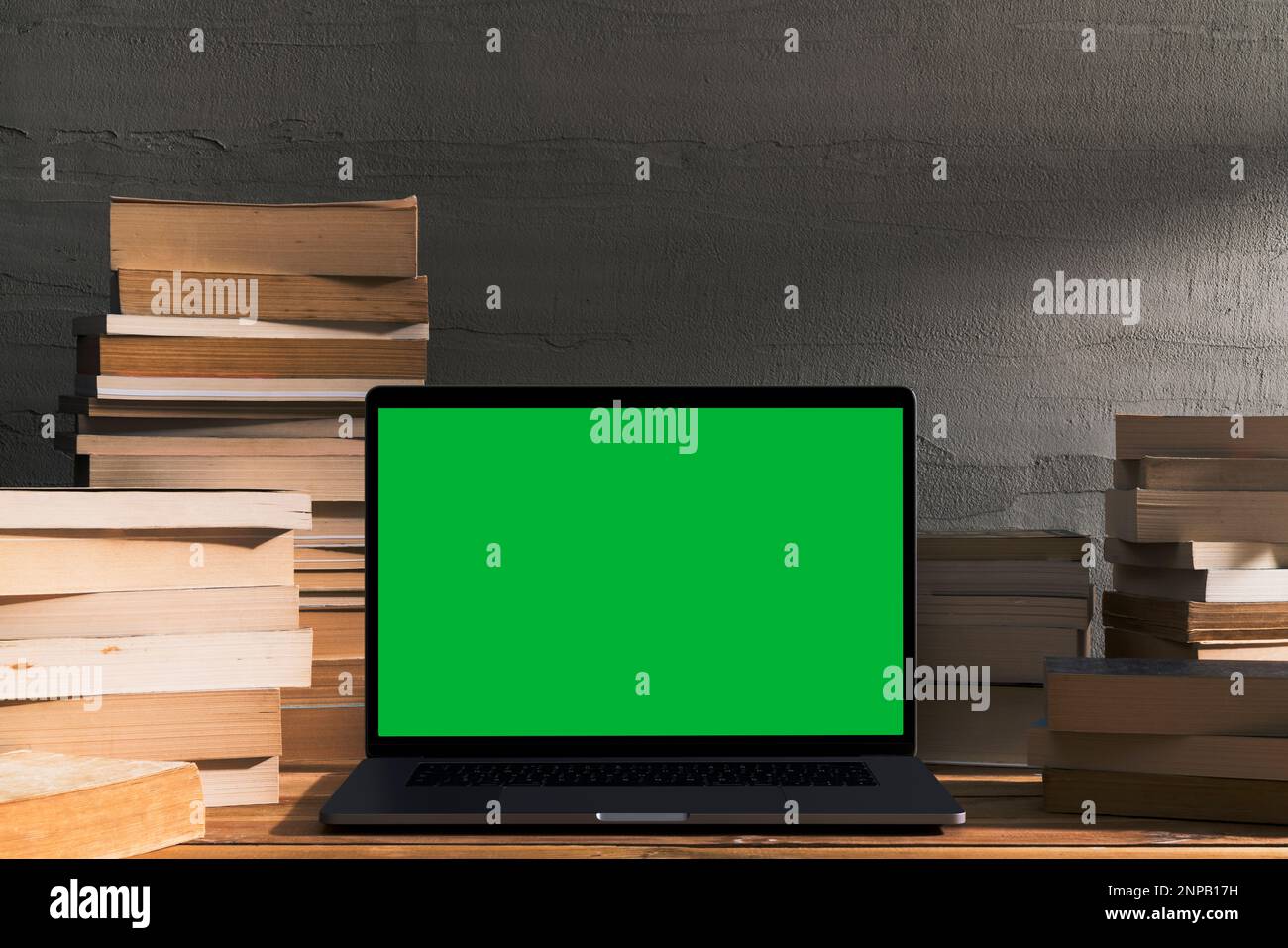 Chroma Key Laptop mit grünem Bildschirm und einem Stapel Bücher auf einem Holztisch vor einer Betonwand. Tischaufnahme des Innenraums mit Fensterlichteffekt Stockfoto