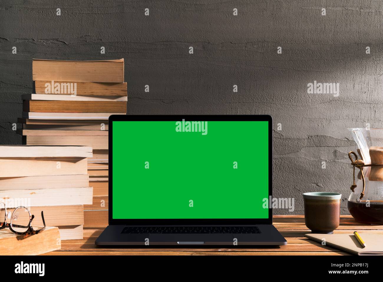 Chroma Key Laptop mit grünem Bildschirm und einem Stapel Bücher auf einem Holztisch vor einer Betonwand. Tischaufnahme des Innenraums mit Fensterlichteffekt Stockfoto