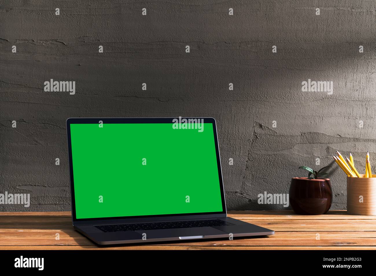 Chroma Key Green Screen, Laptop mit abgewinkelter Sicht auf Holztisch vor Betonwand. Tischaufnahme des Innenraums mit Fensterlichteffekt. Stockfoto
