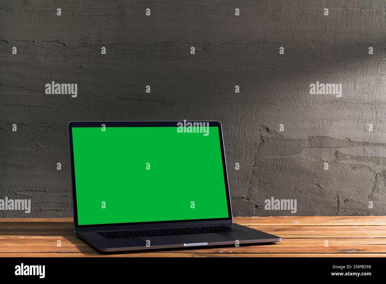 Chroma Key Green Screen, Laptop mit abgewinkelter Sicht auf Holztisch vor Betonwand. Tischaufnahme des Innenraums mit Fensterlichteffekt. Stockfoto