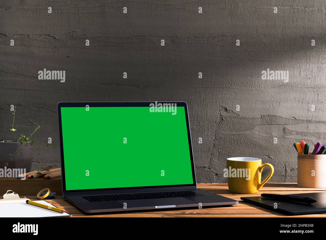 Chroma Key grüner Bildschirm, Laptop mit abgewinkelter Sicht auf Holztisch des Designers vor Betonwand. Tischaufnahme des Innenraums mit Fensterbeleuchtung Stockfoto