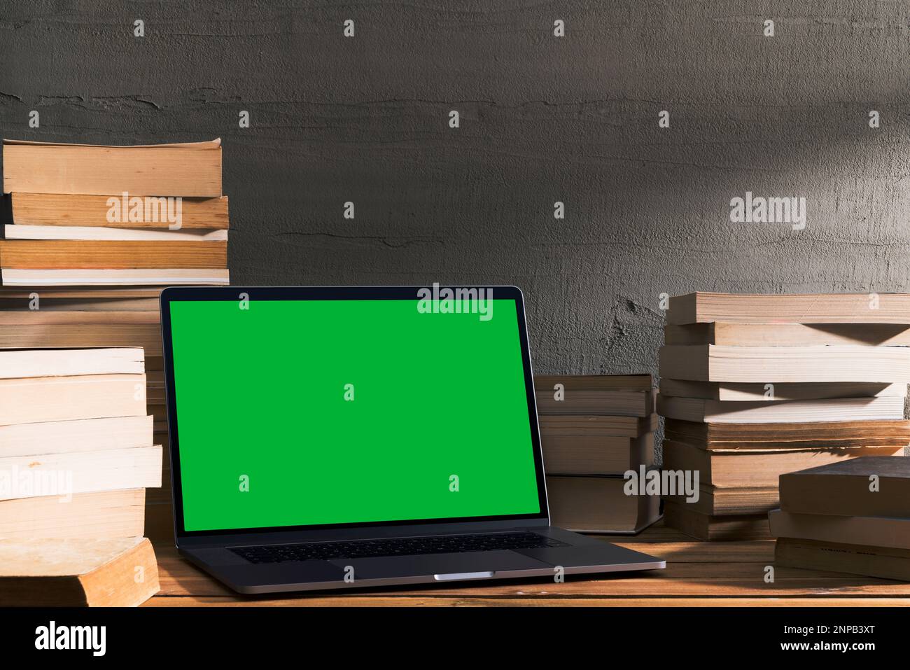 Chroma Key Laptop mit grünem Bildschirm und einem Stapel Bücher auf einem Holztisch vor einer Betonwand. Tischaufnahme des Innenraums mit Fensterlichteffekt Stockfoto