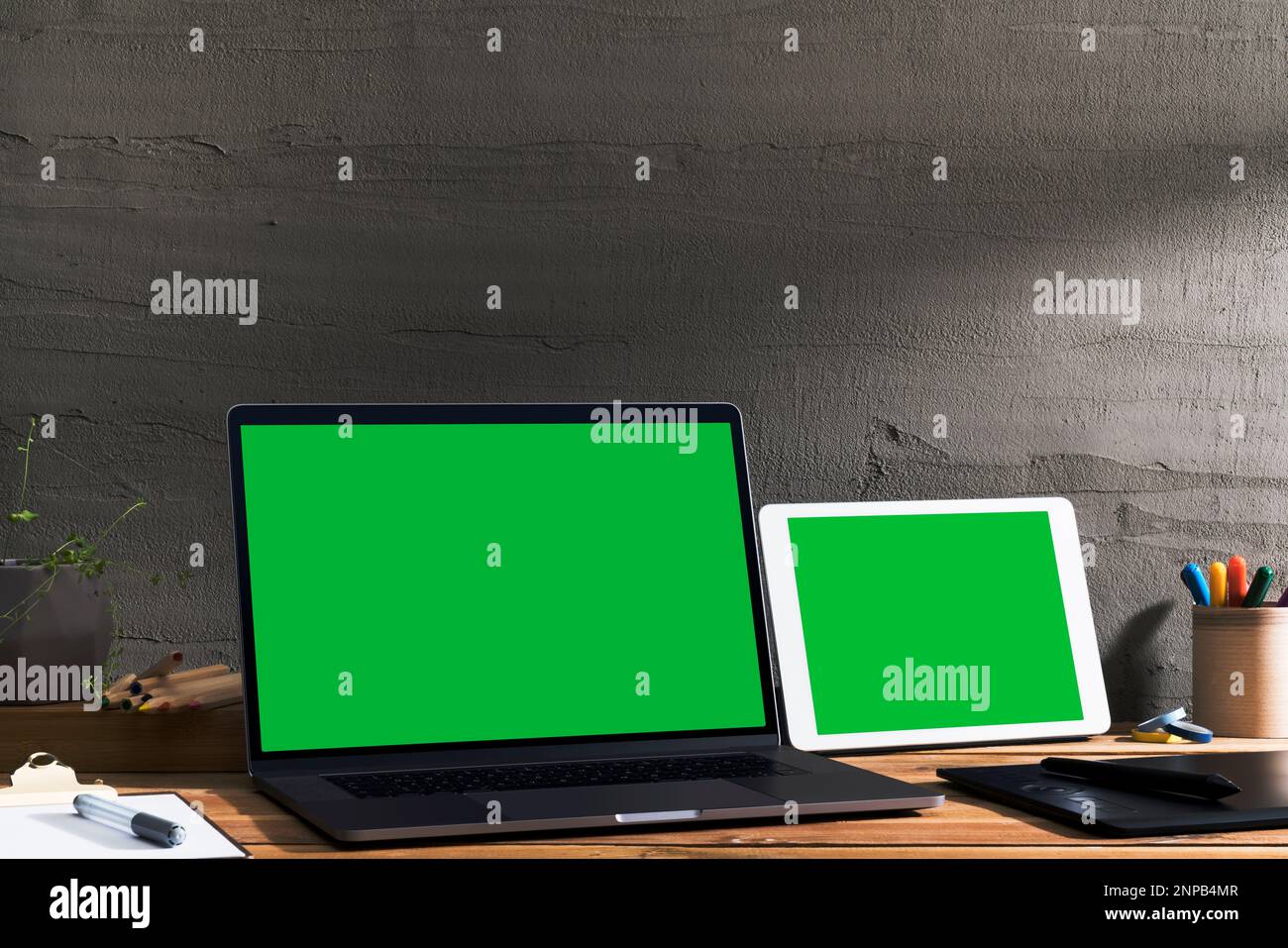 Chroma Key grüner Bildschirm, Laptop mit abgewinkelter Ansicht und digitales Tablet auf Holztisch des Designers vor Betonwand. Tischaufnahme des Innenraums Stockfoto