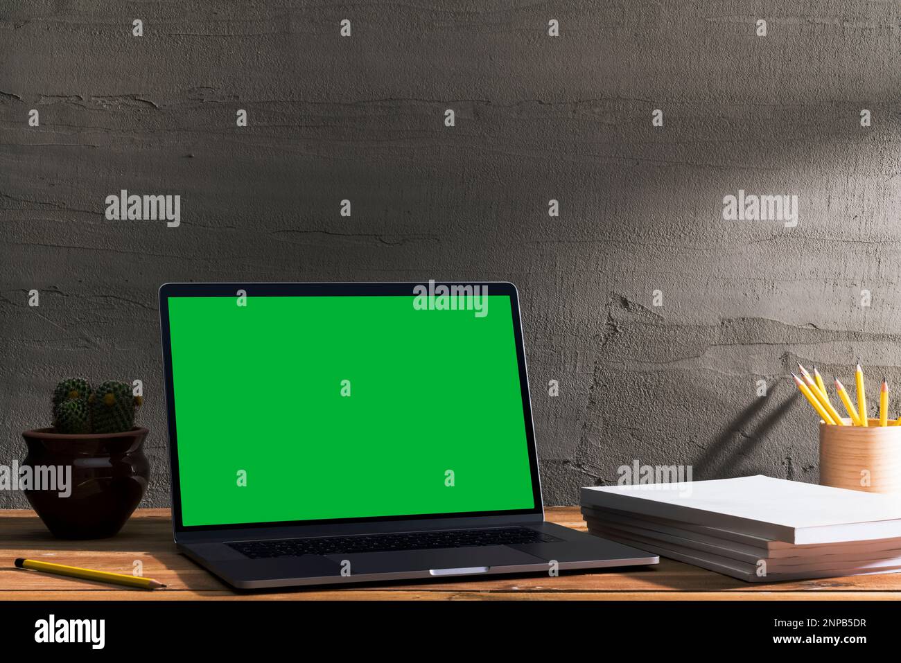 Chroma Key grüner Bildschirm, Laptop mit abgewinkelter Ansicht und weißem Magazinstapel auf Holztisch vor Betonwand. Tischaufnahme des inneren Spaces Stockfoto